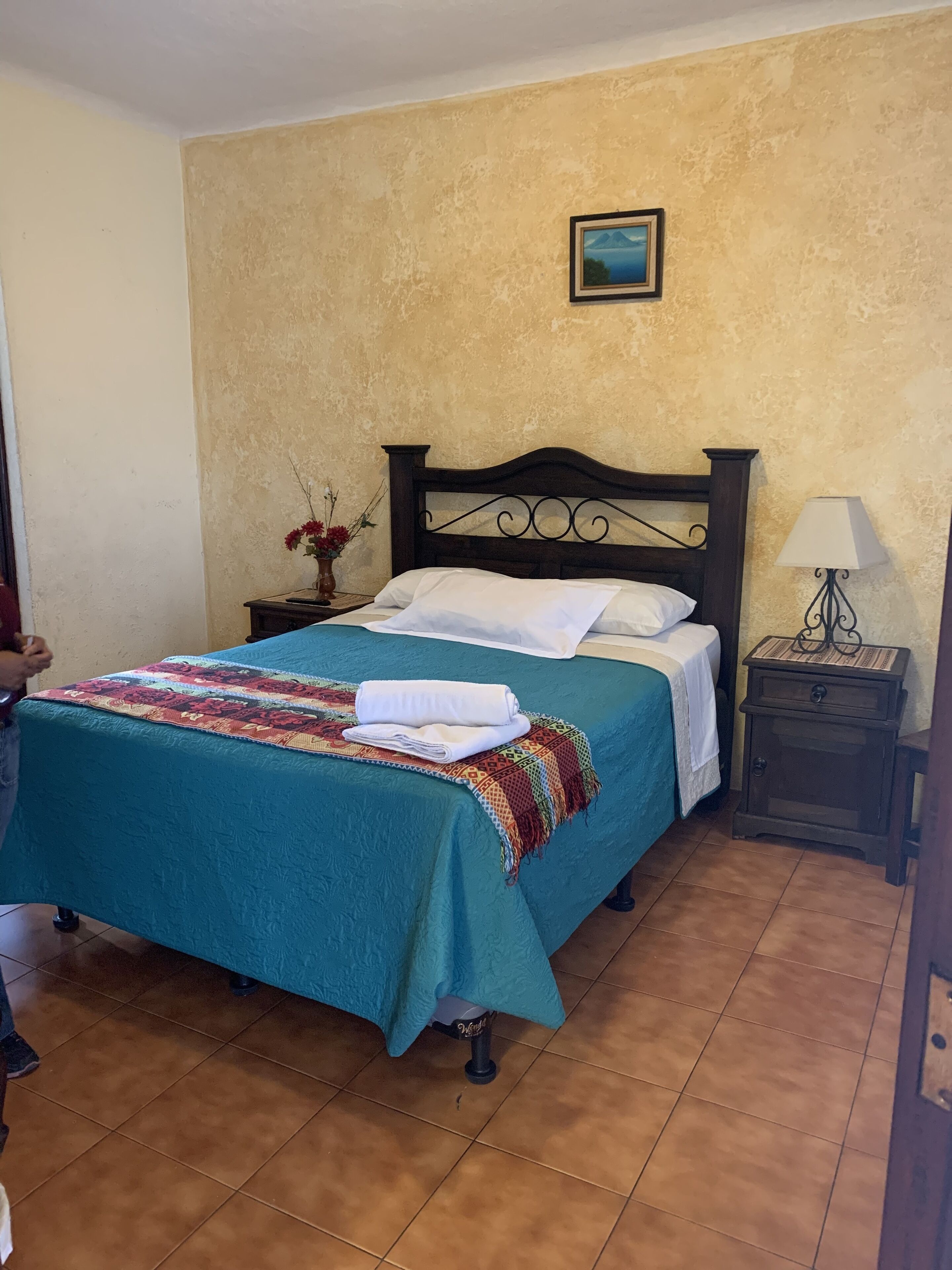 A neatly made bed with a teal bedspread and white linens, a small bedside table with a lamp, and a framed picture on the wall.