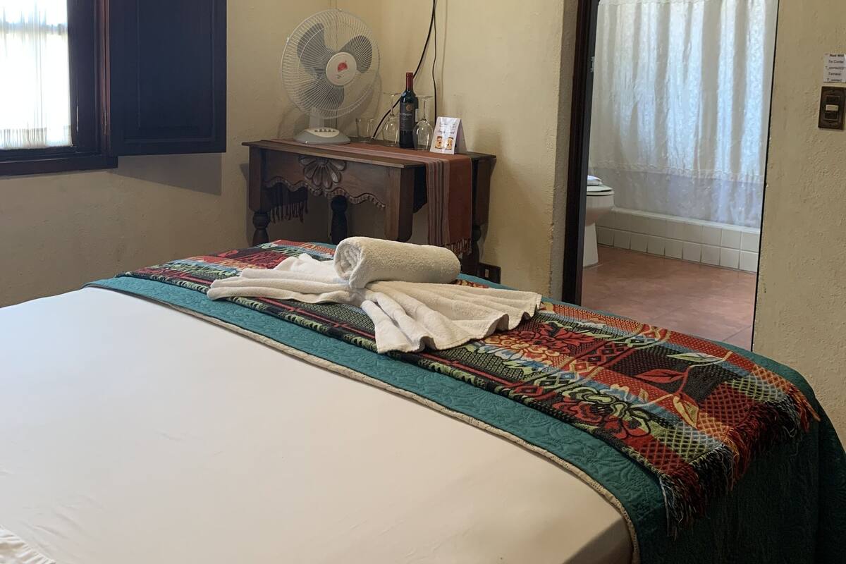 Quarto de hotel com uma cama, mesa de cabeceira com um ventilador, TV fixada na parede e banheiro visível através de uma porta aberta.