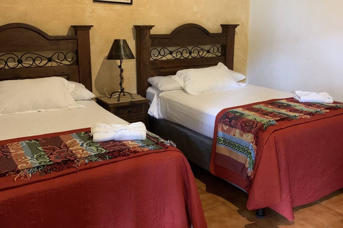 Quarto com duas camas, cada uma com colcha vermelha e travesseiros brancos. Cada cama tem uma cabeceira de madeira e uma pequena mesa com abajur entre elas. As paredes são bege, e há um quadro pendurado na parede acima das camas.