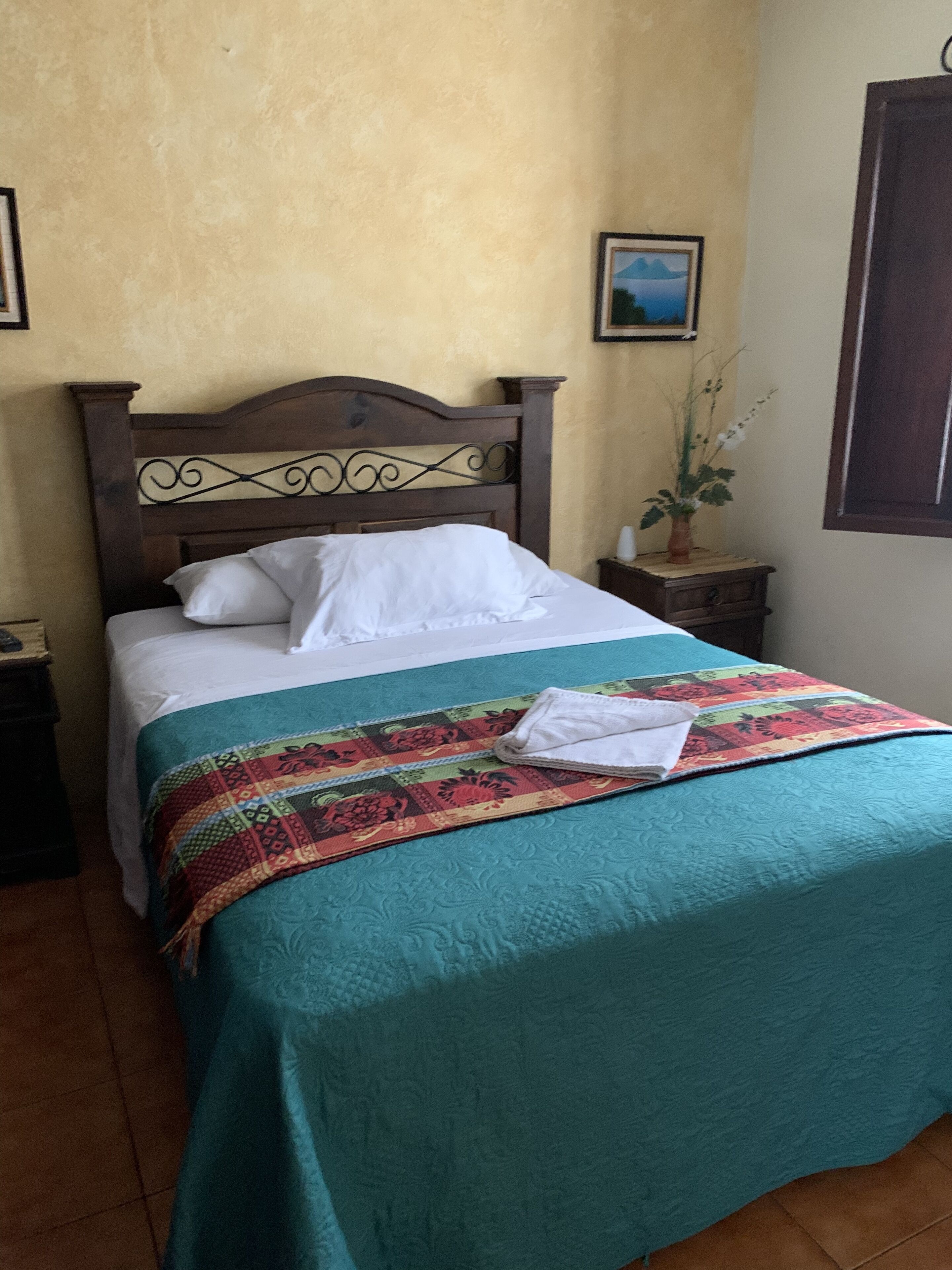 A neatly made bed with a teal bedspread and a colorful quilt, a wooden headboard, and a small bedside table with a plant.