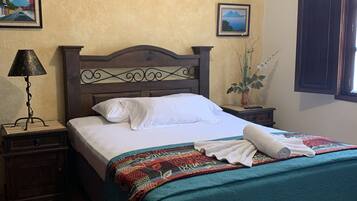 Uma cama bem arrumada com colcha azul-turquesa e almofadas brancas, uma pequena mesa de cabeceira com abajur, e uma janela embutida na parede com venezianas escuras.