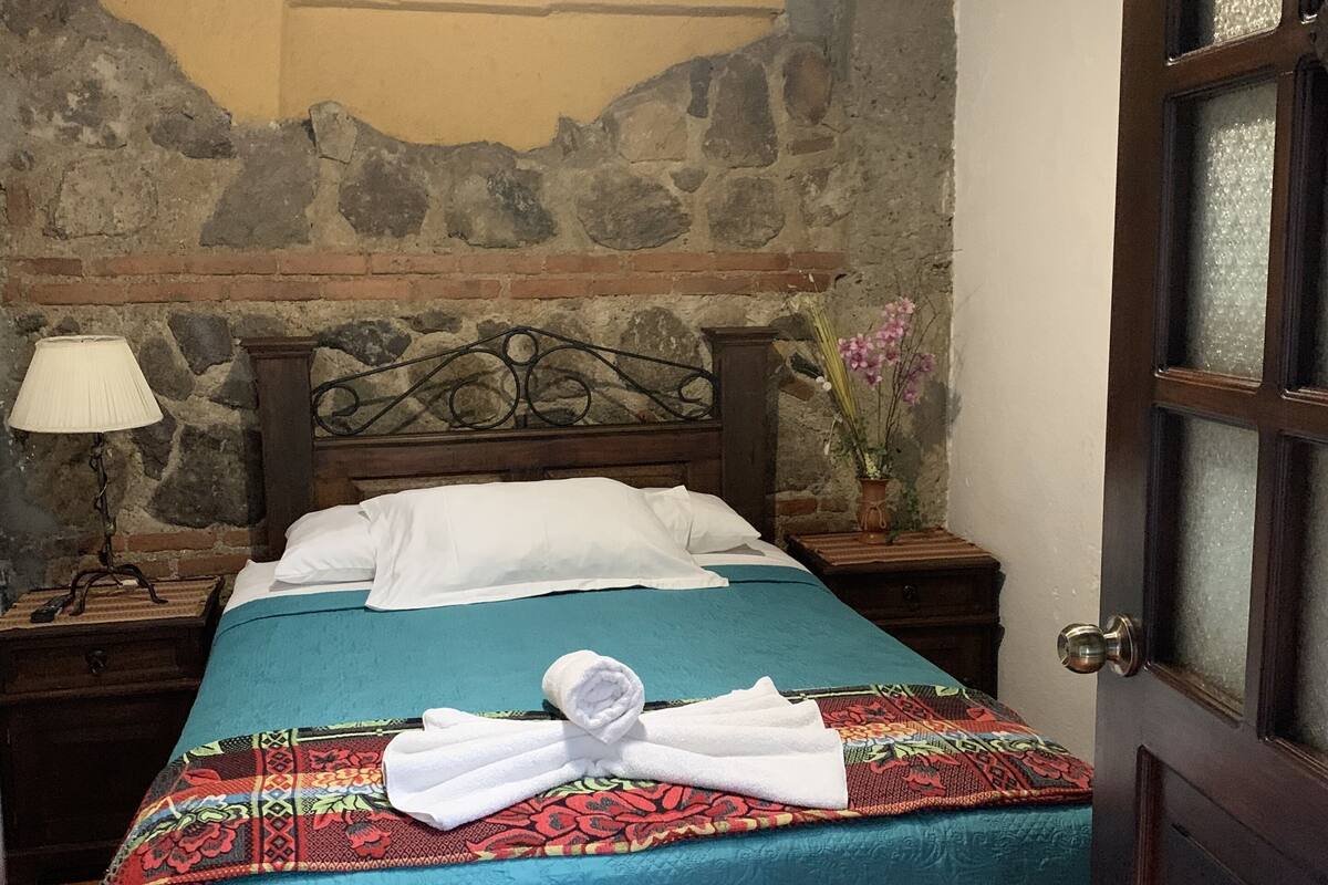 Uma cama bem arrumada com colcha azul e almofadas brancas, cabeceira de madeira, parede de pedra, mesa de cabeceira com abajur e porta com painéis de vidro fosco.