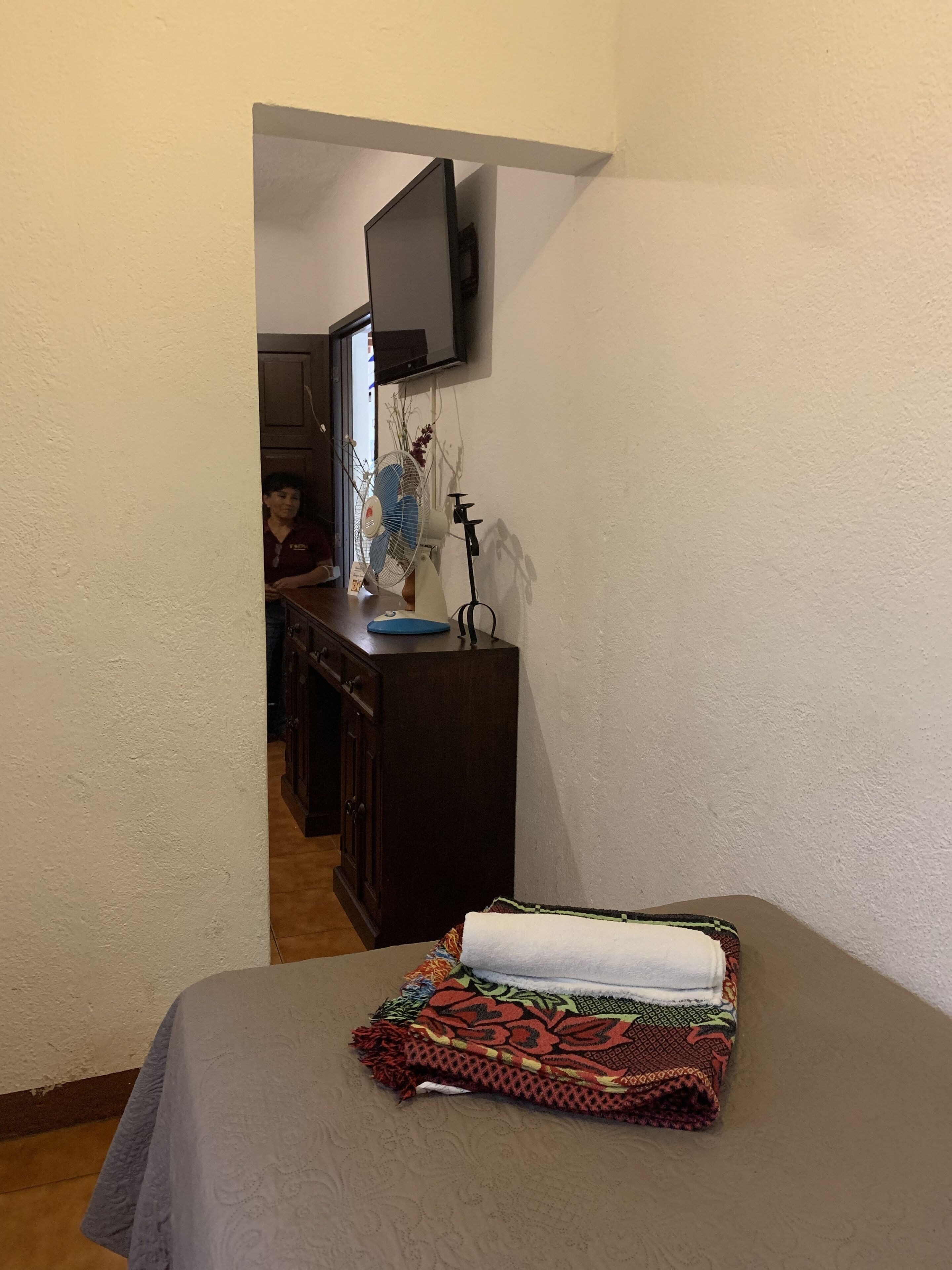 Habitación de hotel con una cama, un televisor montado en la pared, un escritorio con un ventilador y vista a la habitación contigua.