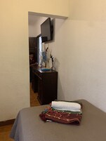 Habitación de hotel con una cama, un televisor montado en la pared, un escritorio con un ventilador y vista a la habitación contigua.