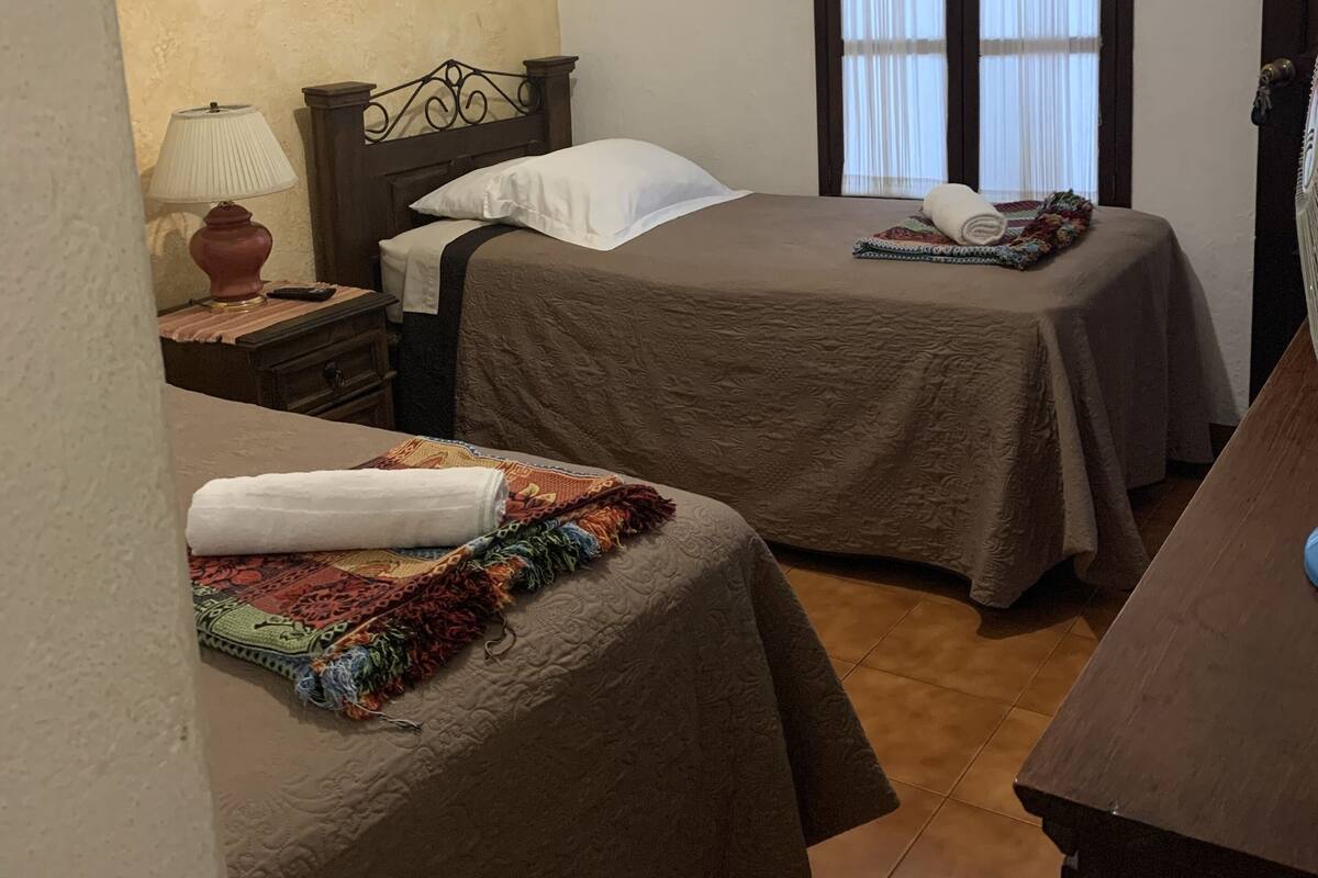 Um quarto pequeno e acolhedor com duas camas, uma mesa de cabeceira, um abajur e uma janela com cortinas.
