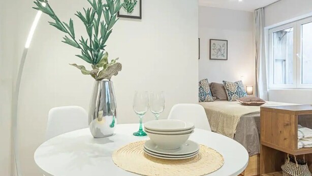 Een eetgedeelte met een ronde tafel, witte stoelen, een vaas met groene planten, en een bed met een witte hoofdbord op de achtergrond.