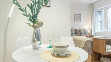 A dining area with a round table, white chairs, a vase with green plants, and a bed with a white headboard in the background.