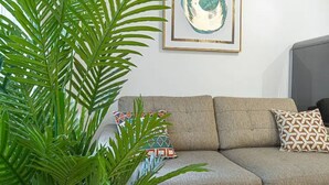 Een zithoek met een grijze bank, decoratieve kussens, een grote groene plant en een kunstwerk in een lijst aan de muur.