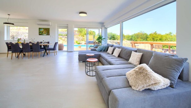 Un vaste espace de vie comprenant un canapé gris, une table à manger avec des chaises, et une vue sur une piscine grâce à de grandes fenêtres.