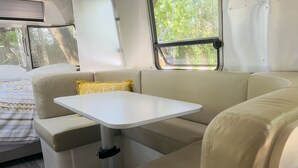 A compact RV interior with a sofa, a small table, and a window providing a view of greenery outside. - Airstream Escape + Pool + Fenced Yard (Austin)