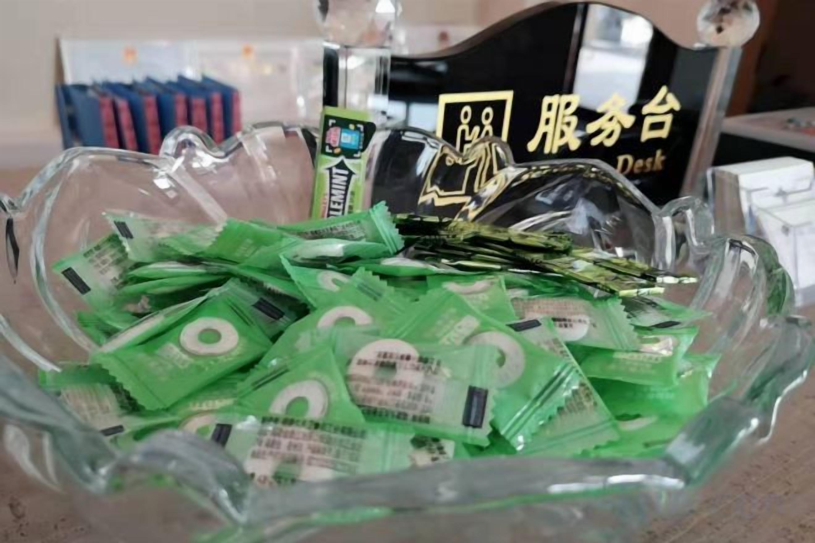 Eine Glasbowl, gefüllt mit grünen Packungen, möglicherweise Snacks oder kleine Gegenstände, auf einem Tisch mit einer Dekoration und einem Schild im Hintergrund.