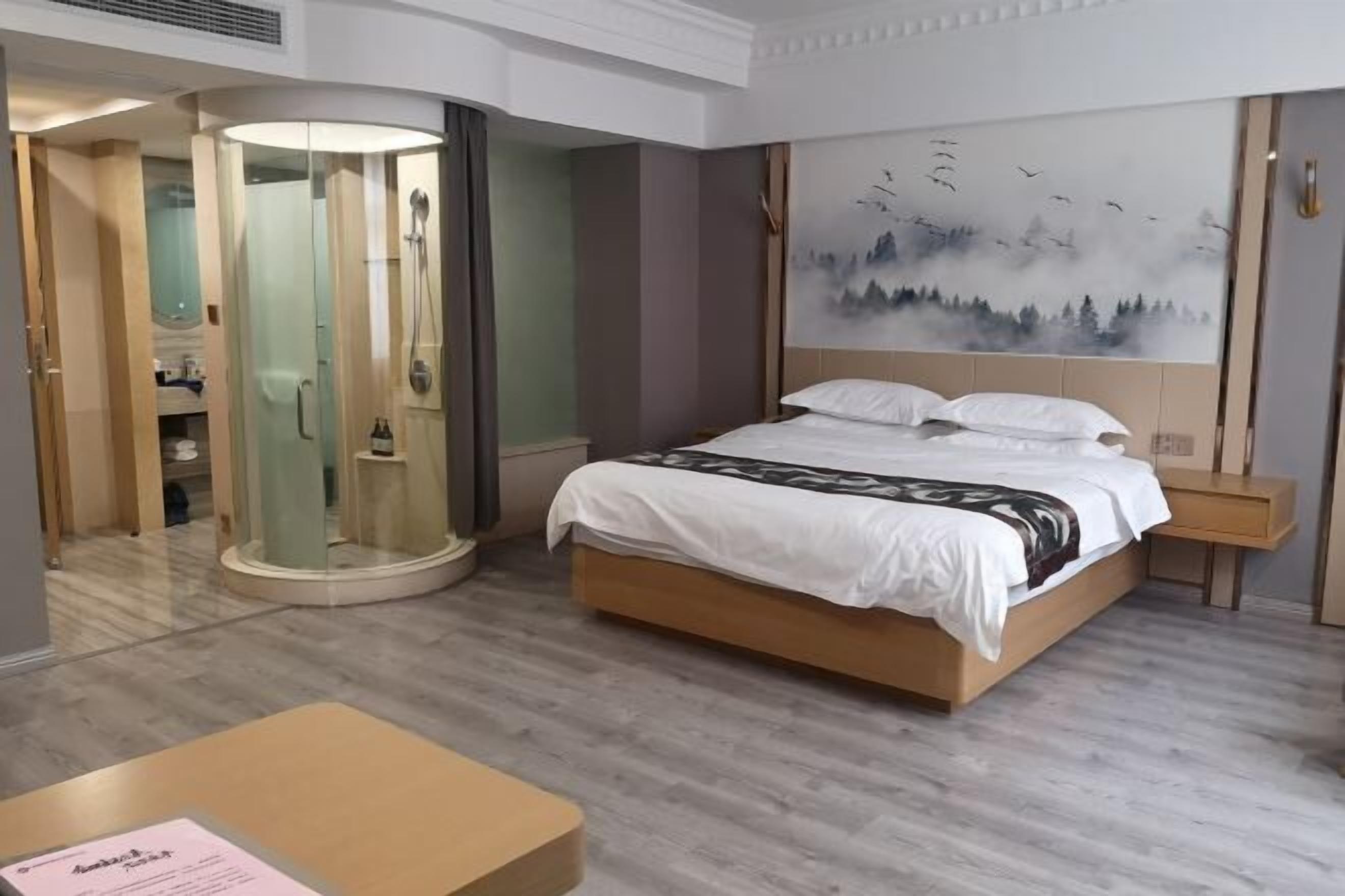 A hotel room with a bed, a glass-enclosed shower, a painting on the wall, and a small table.