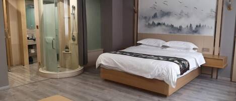 A hotel room with a bed, a glass-enclosed shower, a painting on the wall, and a small table.