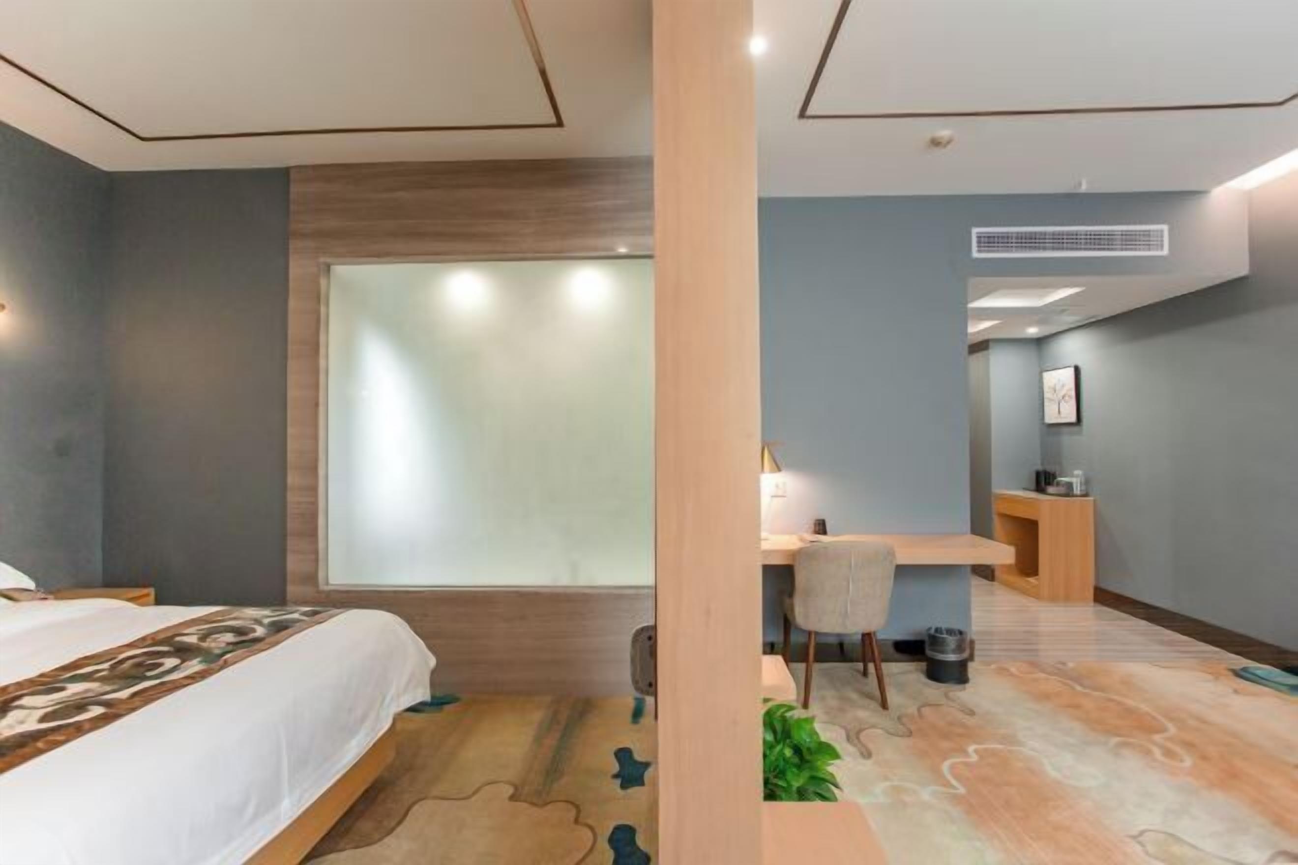 A hotel room with a bed, a desk, a chair, a small plant, and a window with frosted glass.