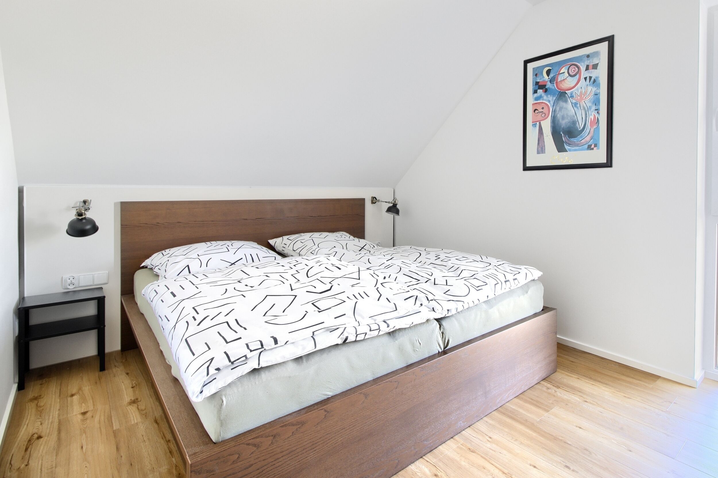 Une chambre à coucher avec un lit, une table de chevet, une lampe fixée au mur et un tableau encadré.