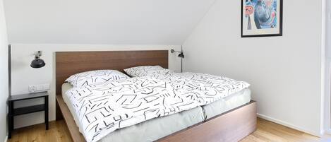 Une chambre à coucher avec un lit, une table de chevet, une lampe fixée au mur et un tableau encadré.