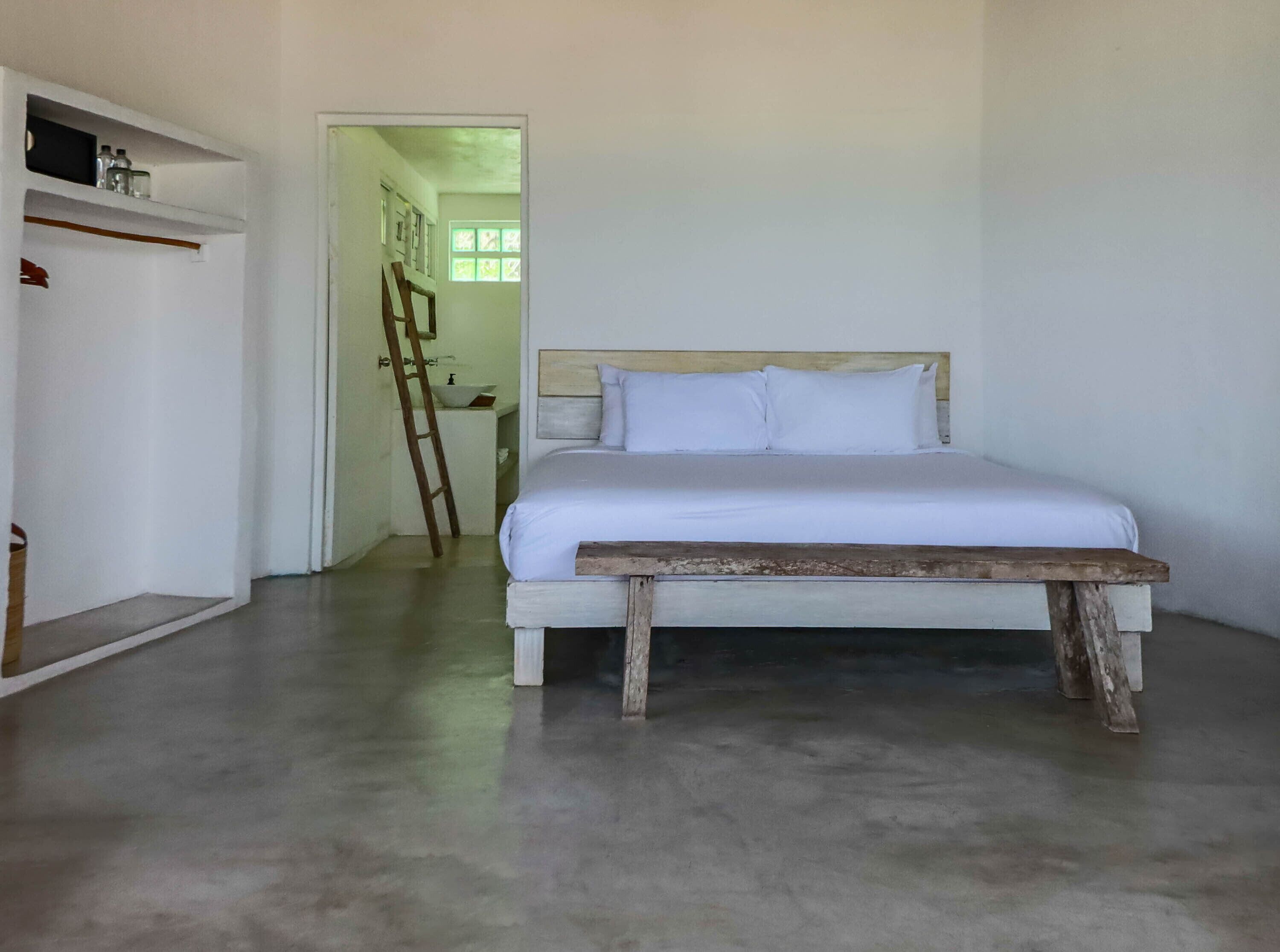 Ein Schlafzimmer mit einem Bett, einer Holzbank, einem Kleiderschrank und einem durchgehenden Türspalt sichtbaren Badezimmer.