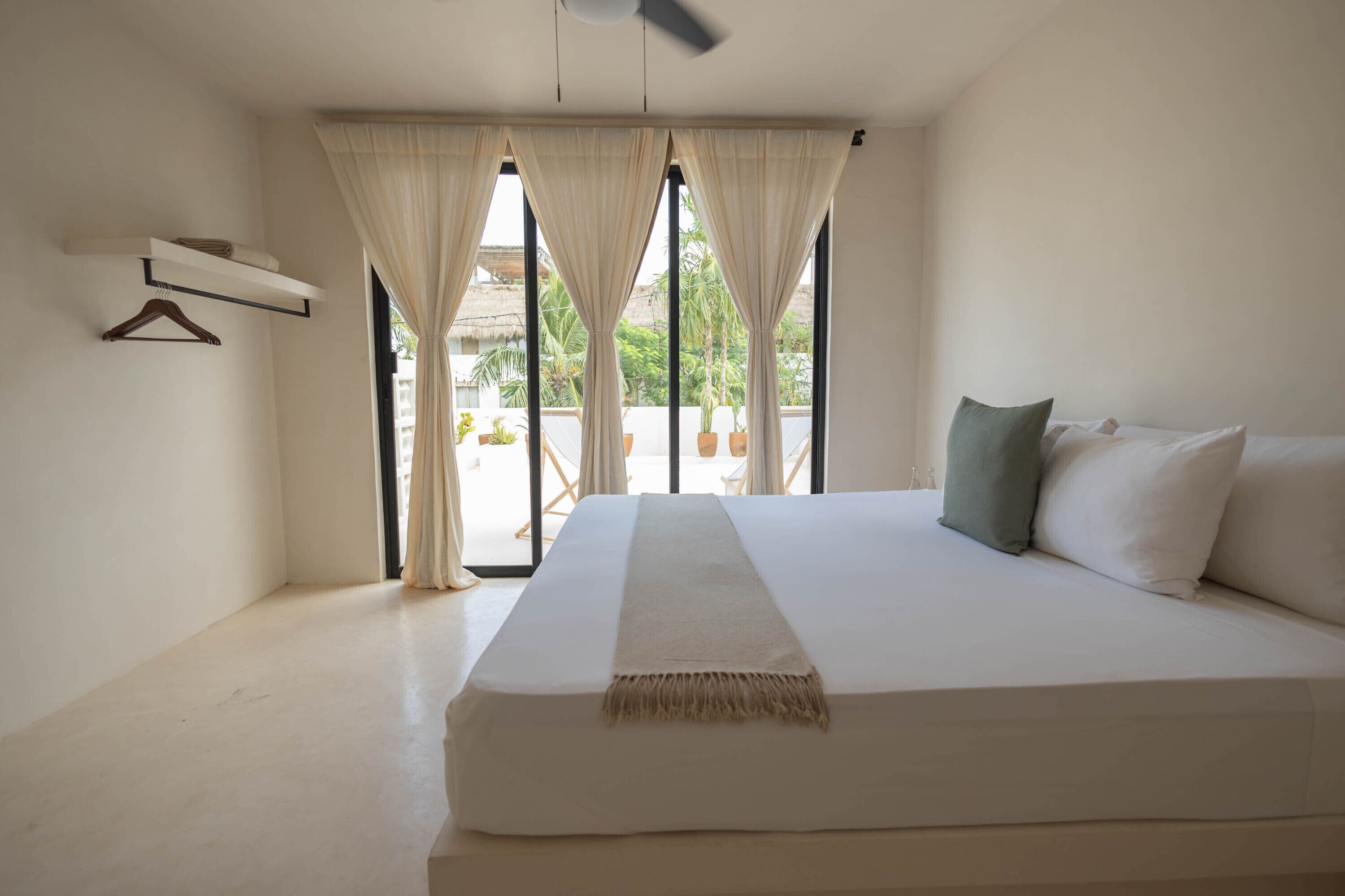 Una cama grande con una colcha blanca y un plaid color beige, un ventilador de techo y vistas al exterior a través de amplias ventanas con cortinas.