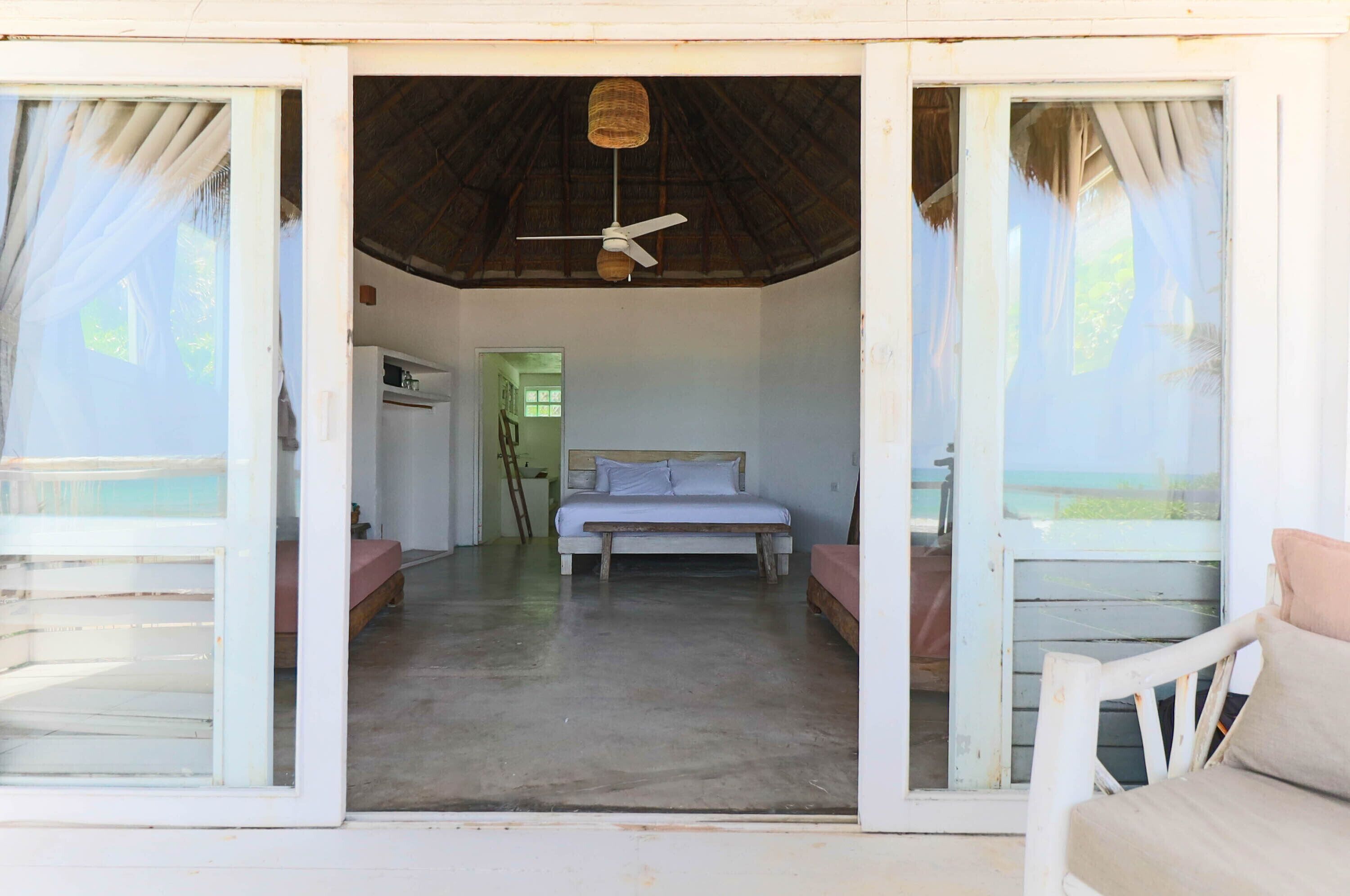 Una habitación con cama, ventilador de techo y vista a la playa por la ventana.
