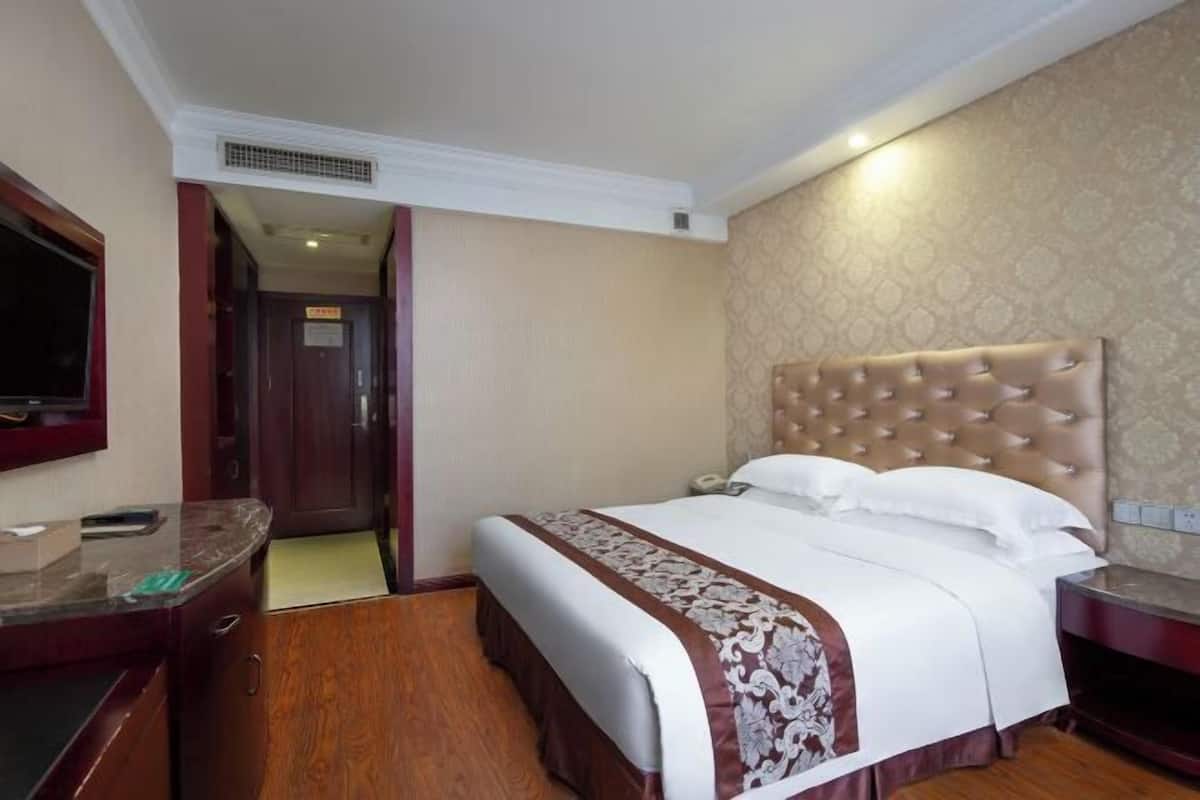 Ein Hotelzimmer mit einem großen Bett, einem Holztisch, einem Fernseher und einer Tür zu einem weiteren Raum.