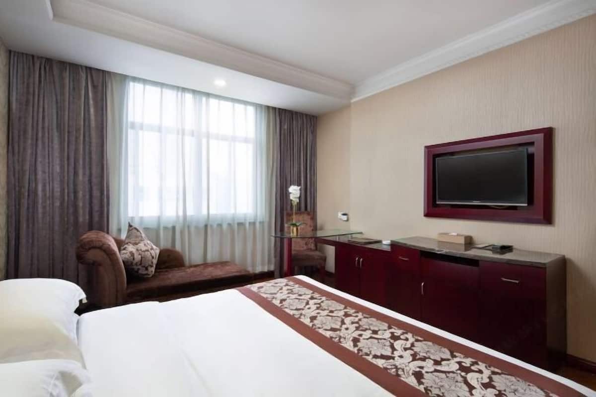 Ein Hotelzimmer mit einem Bett, einem an der Wand befestigten Fernseher, einem Schreibtisch mit Stuhl und einem Fenster mit Vorhängen.