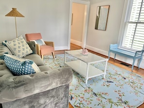 Ein Wohnzimmer mit einem grauen Sofa, einem rosa Sessel, einem weißen Couchtisch und einer blauen Bank an einem Fenster.