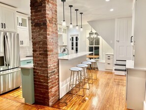 A modern kitchen with a brick column, stainless steel appliances, white cabinetry, and a wooden floor. - Large newly renovated Cape Victorian 5 blocks from the beach, sleeps 12 (Cape Charles)