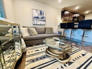A modern living room with a glass coffee table, a grey sofa, and a patterned rug.