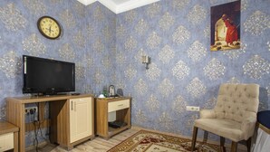 A room with a television on a wooden cabinet, a beige armchair, a patterned carpet, and a wall clock. - GLOBAL OTEL (Mus)