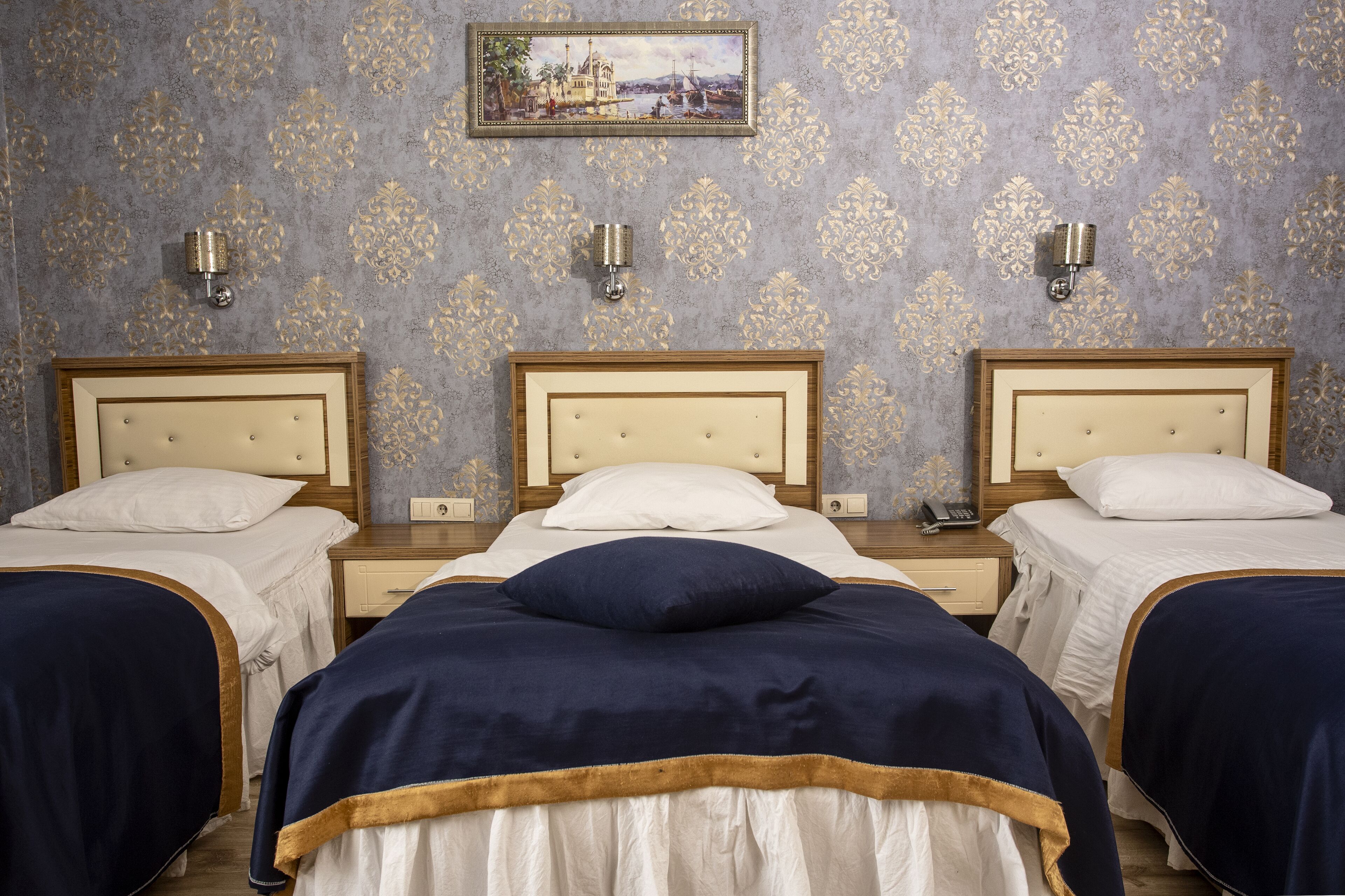 Habitación de hotel con tres camas, cada una con cabecero de madera y ropa de cama blanca. Hay dos mesitas de noche con lámparas y un pequeño cuadro en la pared sobre la cama central.