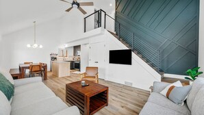 Uma sala de estar moderna com sofá, mesa de centro e área de jantar. Uma escadaria leva a um andar superior com varanda.