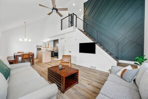 A modern living room with a sofa, coffee table, and a dining area. A staircase leads to an upper level with a balcony. - Locally Owned 4BR w/Hot Tub (Traverse City)