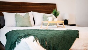 Cama bem arrumada com manta verde, mesa de cabeceira de madeira com caneca branca e uma pequena planta, e abajur branco no criado-mudo.