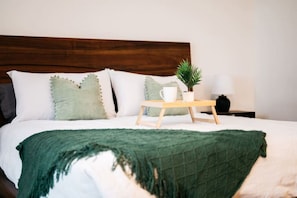 A neatly made bed with a green throw blanket, a wooden bedside table with a white mug and a small plant, and a white lamp on the nightstand. - Locally Owned 4BR w/Hot Tub (Traverse City)