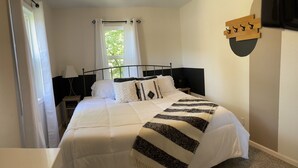 A neatly made bed with white bedding and a black and white striped blanket, a bedside table with a lamp, a window with sheer curtains, and a wall-mounted coat rack. - The Capitol House-East Village/Downtown, King Bed (Des Moines)