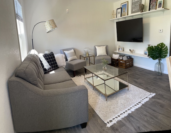 A modern living room with a grey sofa, a glass coffee table, and a flat-screen TV mounted on the wall.