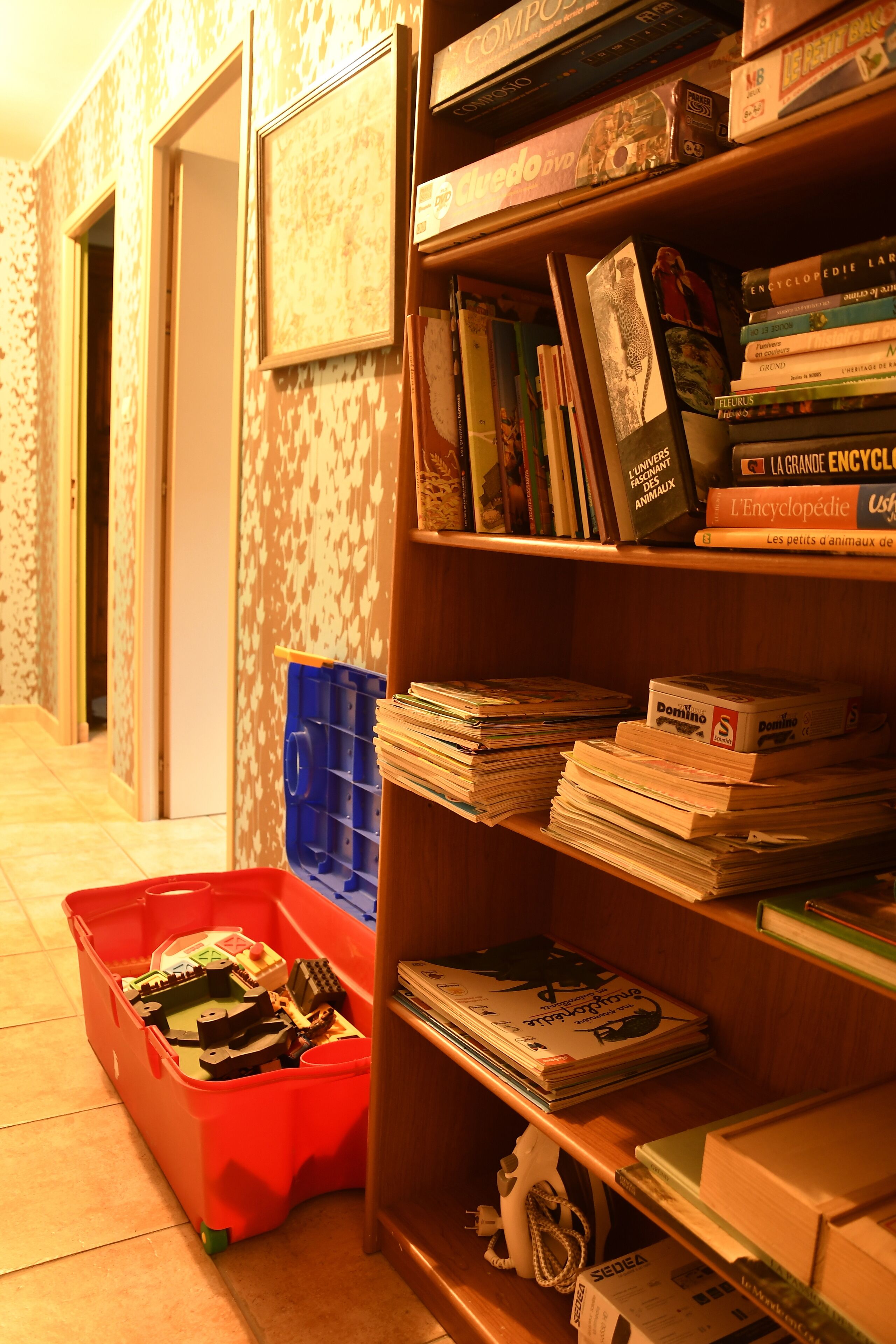 A room with a bookshelf filled with books and a red storage bin with toys.