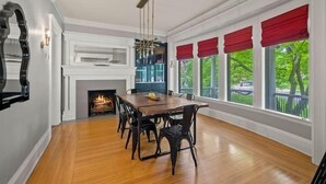 A dining room with a wooden table and chairs, a fireplace, and large windows with red valances.
