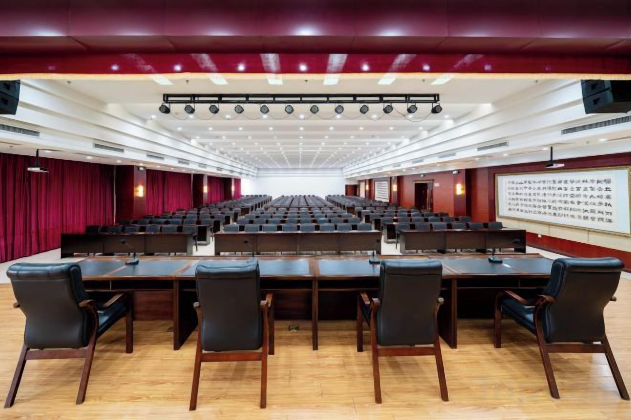 Un salón de conferencias con mesas y sillas dispuestas en filas, un escenario con una pantalla y un telón de fondo rojo.