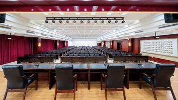 Un salón de conferencias con mesas y sillas dispuestas en filas, un escenario con una pantalla y un telón de fondo rojo.