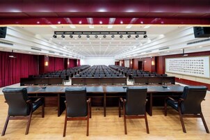 Un salón de conferencias con mesas y sillas dispuestas en filas, un escenario con una pantalla y un telón de fondo rojo.