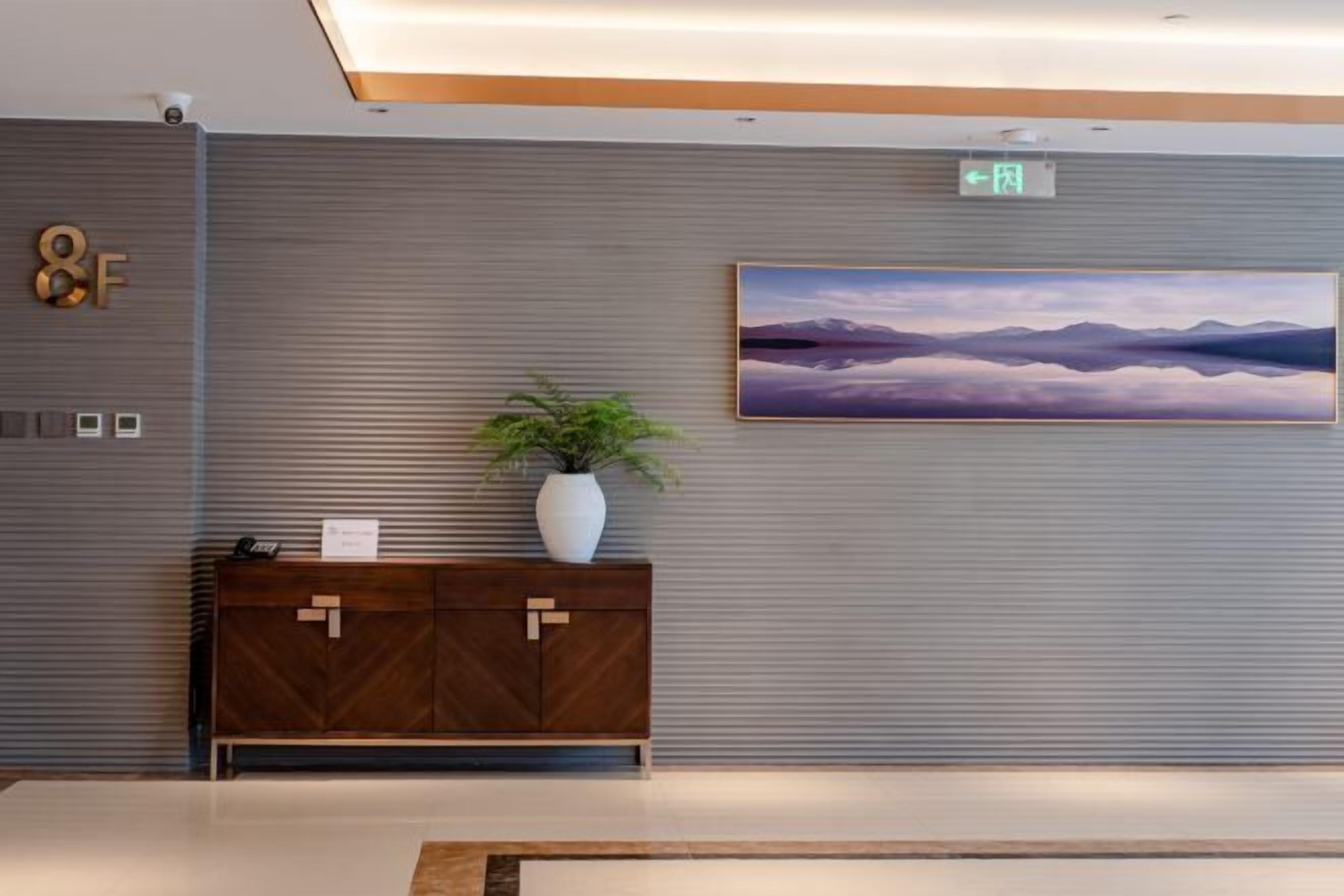 A modern hotel lobby with a large painting of mountains, a wooden cabinet with a plant, and a sign indicating an exit.