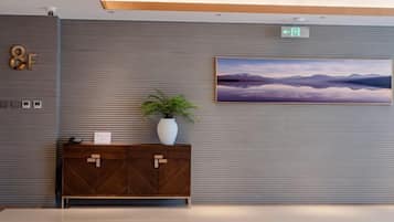 A modern hotel lobby with a large painting of mountains, a wooden cabinet with a plant, and a sign indicating an exit.
