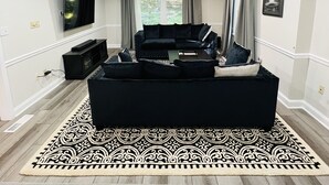 Una sala moderna con un amplio sofá seccional negro, un televisor de pantalla plana montado en la pared y un ventanal con cortinas.