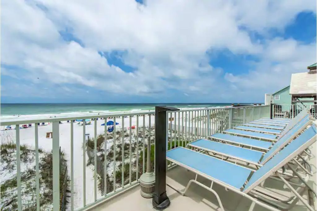 A balcony with sun loungers overlooking a beach with a fence and beach umbrellas.