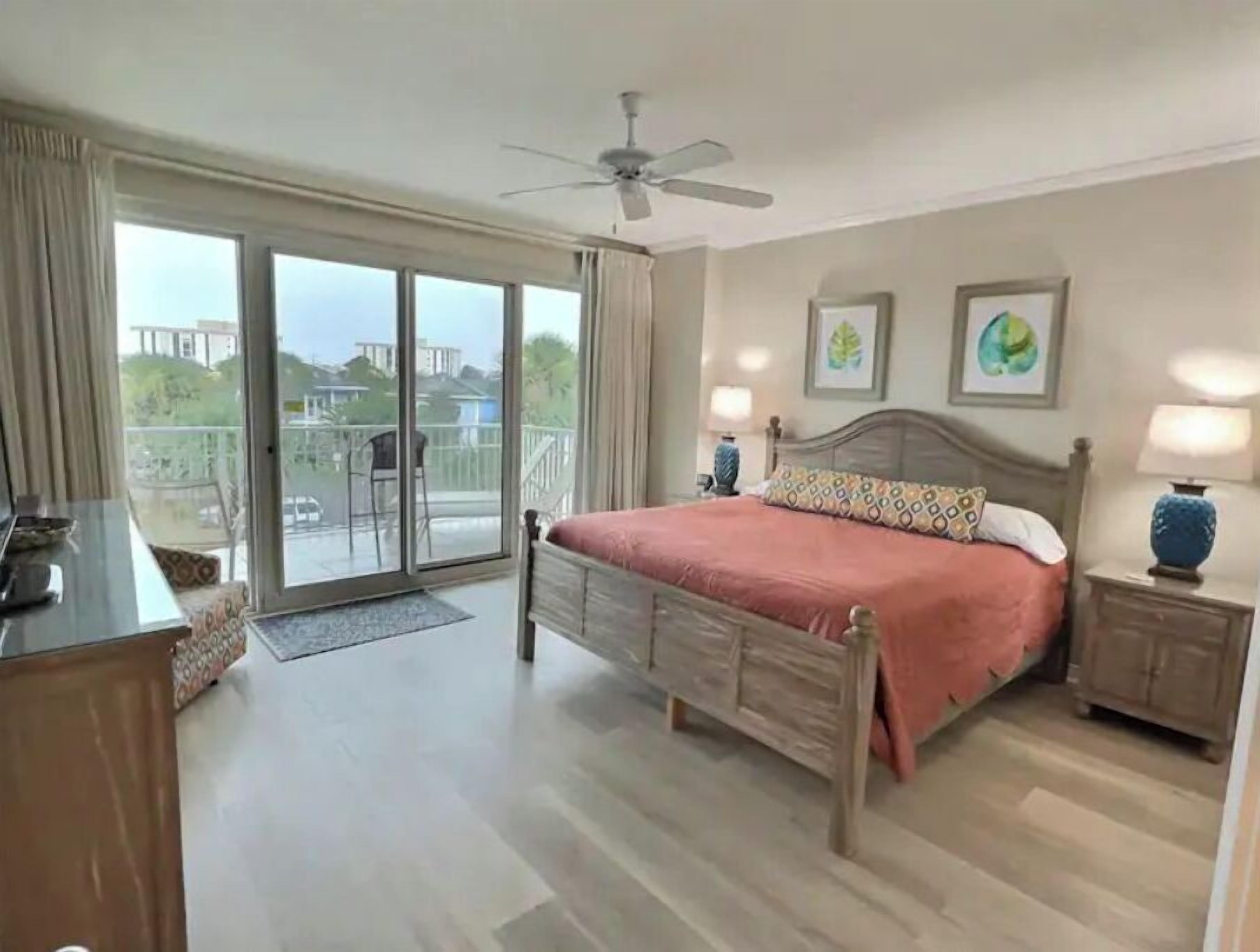 Ein Schlafzimmer mit Bett, Deckenventilator, Nachttisch, Lampe und Blick durch eine Schiebetür auf einen Außenbereich.