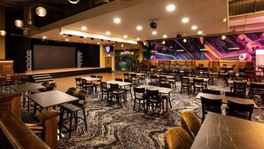 Um espaço amplo e interno com várias mesas e cadeiras, um palco com uma tela grande e um mural vibrante na parede.