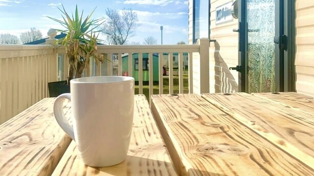 Uma mesa de madeira com uma caneca branca, uma porta de metal com painel de vidro e um céu claro com nuvens.