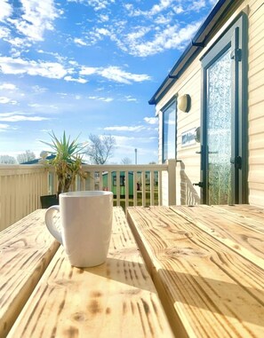 A wooden table with a white mug, a metal door with a glass panel, and a clear sky with clouds. - P27 3 Bedroom Caravan, home from home at Sand le Mere (Hull)