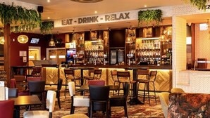 Uma área de bar com um letreiro que diz "EAT - DRINK - RELAX". Há vários espaços de estar com cadeiras e mesas.